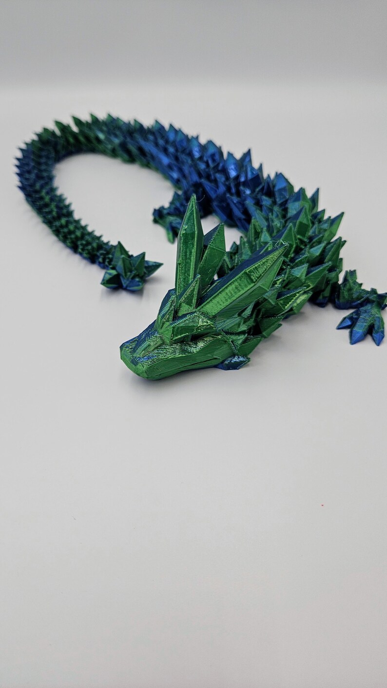 May include: A 3D printed dragon figurine with a green and blue iridescent finish. The dragon is curled up in a defensive posture with its head resting on its tail.
