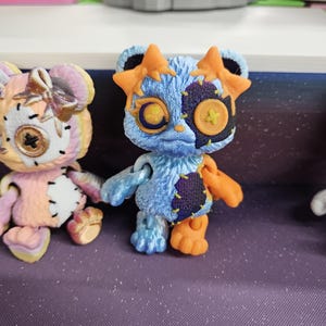 Puede incluir: Primer plano de dos figuras de oso hechas a mano. El oso azul tiene detalles en naranja, un parche cosido y orejas en forma de estrella. El otro oso es rosa y amarillo con un lazo. Ambos tienen ojos de bot&oacute;n y detalles cosidos. Est&aacute;n sobre una superficie p&uacute;rpura.