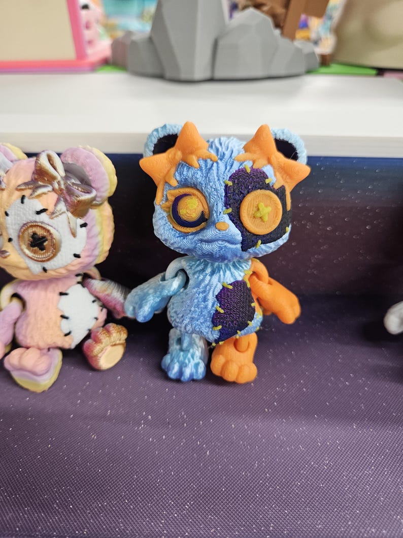 Puede incluir: Primer plano de dos figuras de oso hechas a mano. El oso azul tiene detalles en naranja, un parche cosido y orejas en forma de estrella. El otro oso es rosa y amarillo con un lazo. Ambos tienen ojos de bot&oacute;n y detalles cosidos. Est&aacute;n sobre una superficie p&uacute;rpura.
