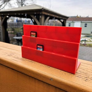 May include: Red plastic game card holder with two game cards inserted. The holder has three slots for game cards. The holder is sitting on a wooden surface.