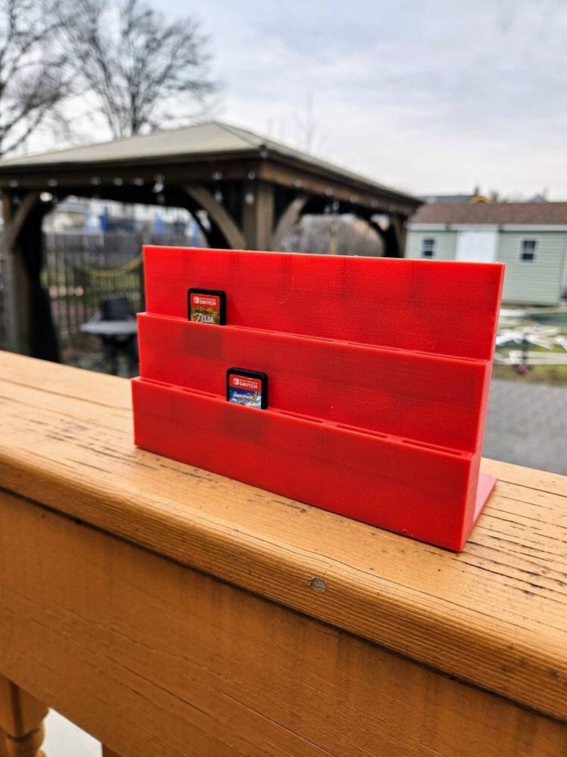 May include: Red plastic game card holder with two game cards inserted. The holder has three slots for game cards. The holder is sitting on a wooden surface.