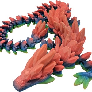 K&ouml;nnte beinhalten: Eine 3D-gedruckte Drachenfigur mit einem Farbverlauf in Rot, Blau und Gr&uuml;n. Der Drache liegt in einer schlafenden Position zusammengerollt, mit dem Kopf auf seinen Vorderpfoten.