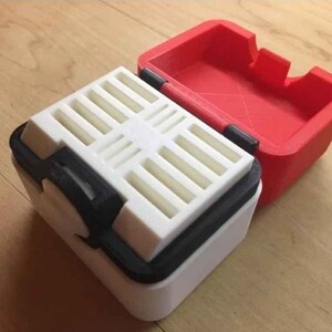 May include: A white and black storage box with a red lid. The box has a grid pattern on the inside and a small knob on the side.