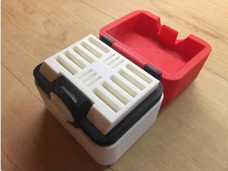 May include: A white and black storage box with a red lid. The box has a grid pattern on the inside and a small knob on the side.