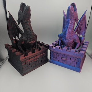 May include: Two dragon sculptures, one black and red, the other blue and purple. Each dragon is perched atop a castle-like structure with open mouths and outstretched wings. The sculptures are detailed and have a fantasy theme.