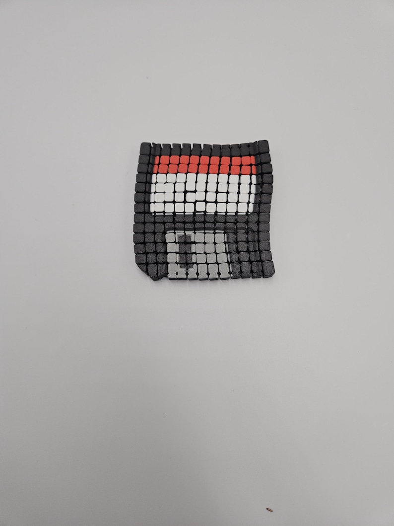 May include: A pixelated floppy disk design in black, red, and white. The square-shaped disk has a grid-like pattern, with a red top border and a white and black center. The design is on a plain white background.