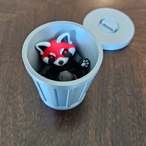 May include: A 3D-printed red panda figurine inside a small, gray trash can with a matching lid. The red panda has a red head, white face, and black body. The trash can is cylindrical with vertical lines.