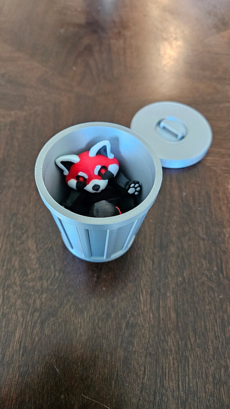 May include: A 3D-printed red panda figurine inside a small, gray trash can with a matching lid. The red panda has a red head, white face, and black body. The trash can is cylindrical with vertical lines.
