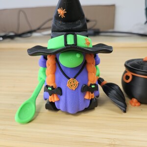 Puede incluir: Una caprichosa figura de gnomo de Halloween, con un sombrero de bruja negro con acentos de ara&ntilde;a naranja, una cara verde y trenzas naranjas. El gnomo lleva un vestido morado y sostiene una cuchara verde. Tambi&eacute;n hay un caldero negro y una escoba.