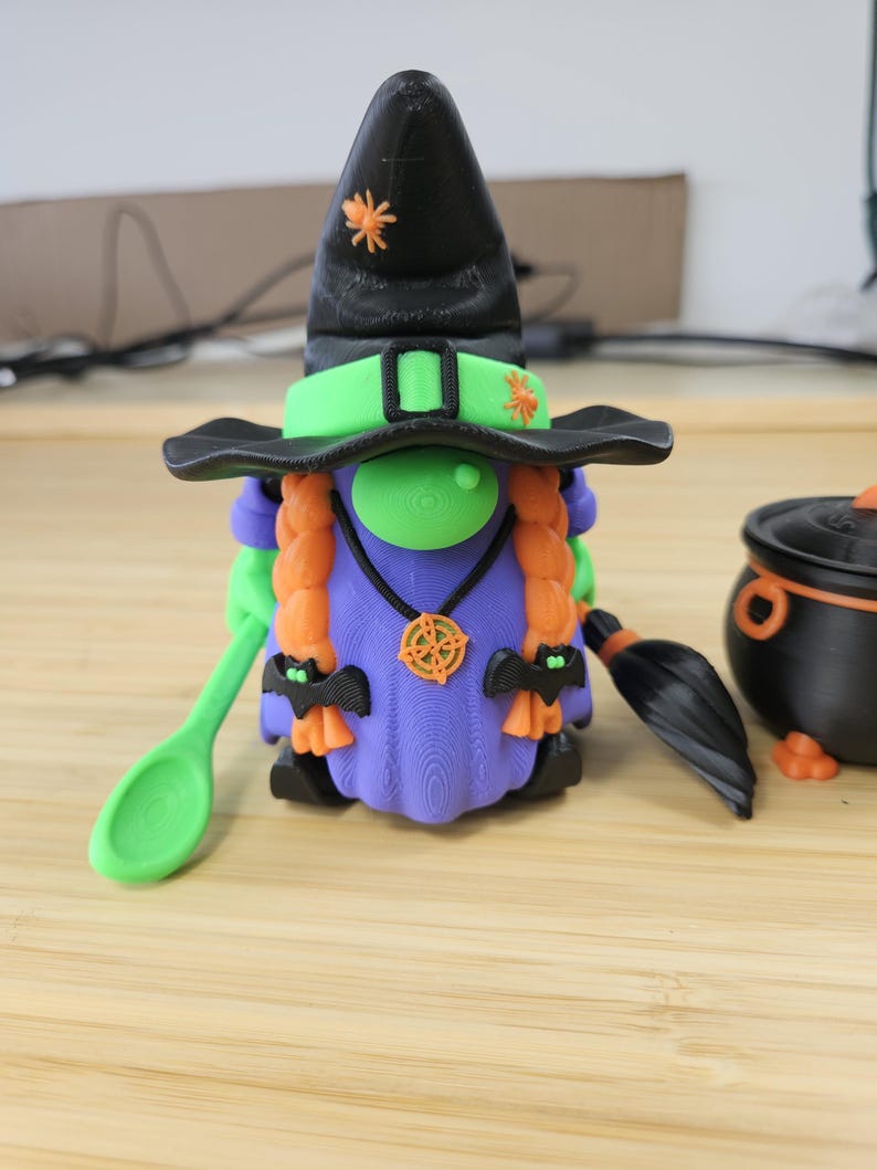 Puede incluir: Una caprichosa figura de gnomo de Halloween, con un sombrero de bruja negro con acentos de ara&ntilde;a naranja, una cara verde y trenzas naranjas. El gnomo lleva un vestido morado y sostiene una cuchara verde. Tambi&eacute;n hay un caldero negro y una escoba.