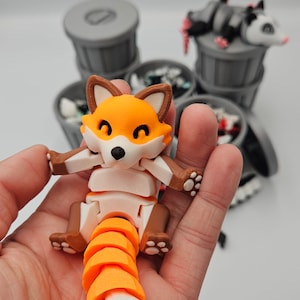 May include: A 3D-printed articulated fox toy in orange, brown, and white, held in a hand. The fox has a segmented tail and is posed with its arms outstretched. Other trash can toys are in the background.