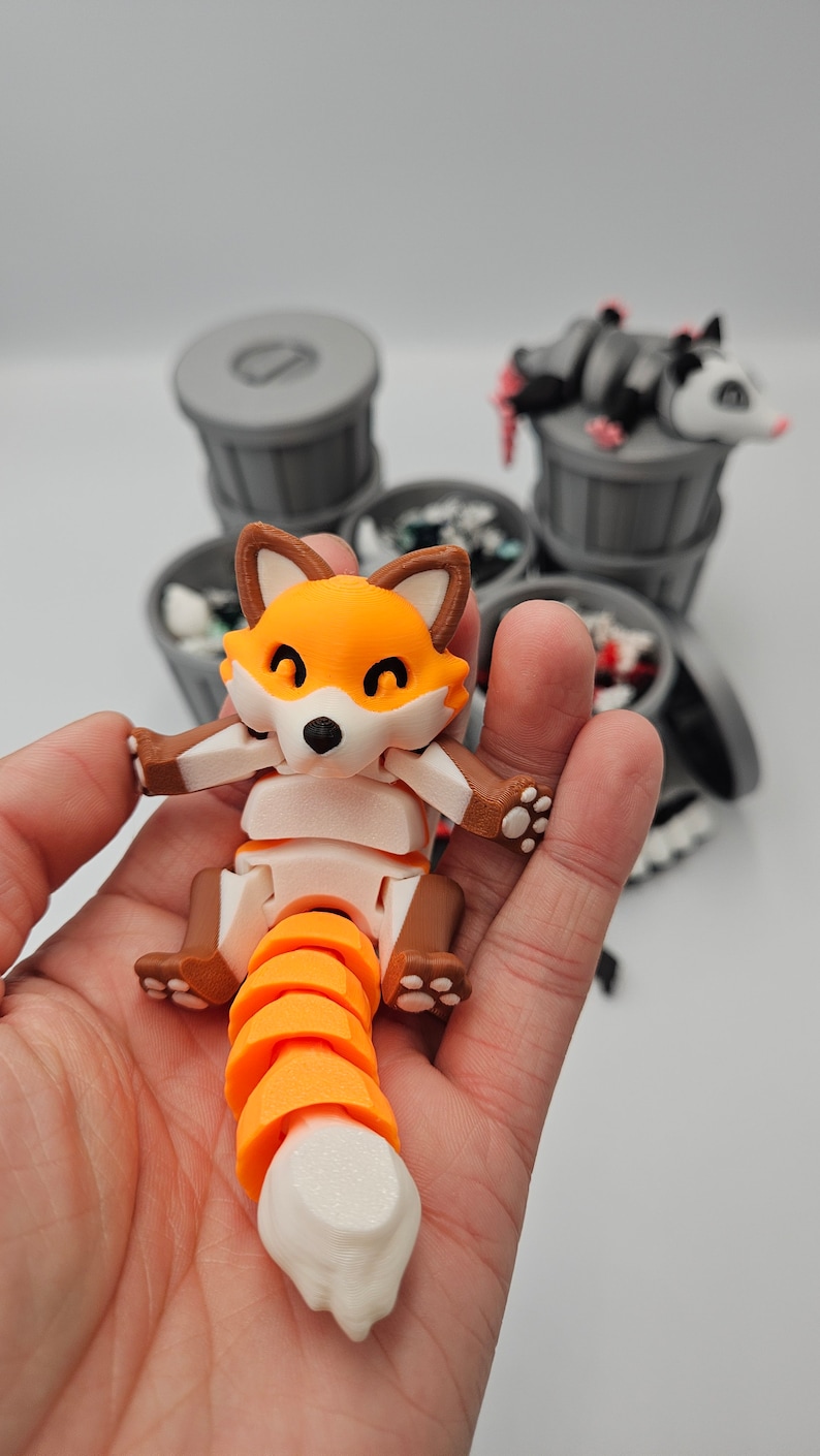 May include: A 3D-printed articulated fox toy in orange, brown, and white, held in a hand. The fox has a segmented tail and is posed with its arms outstretched. Other trash can toys are in the background.