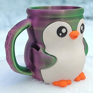May include: A 3D-printed mug shaped like a cartoon penguin. The mug features a white body, black eyes, an orange beak and feet, and a purple and green gradient on the handle and rim. The mug is sitting on a snowy surface.