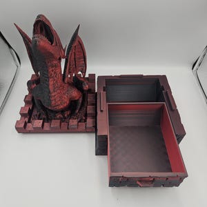 May include: A red and black dragon-themed box with an open compartment. The dragon is perched atop a brick-like base, with its mouth open and wings spread. The box is open, revealing a dark interior.