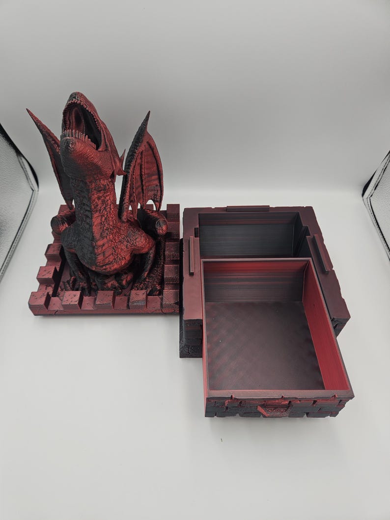 May include: A red and black dragon-themed box with an open compartment. The dragon is perched atop a brick-like base, with its mouth open and wings spread. The box is open, revealing a dark interior.