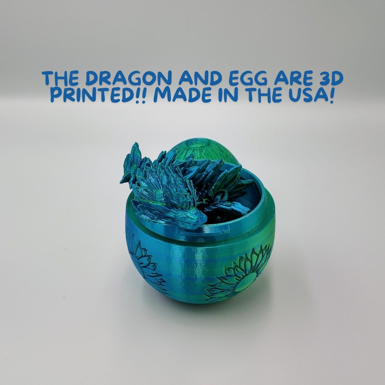 Puede incluir: Una figura de drag&oacute;n impresa en 3D dentro de un recipiente en forma de huevo de color verde azulado con un dise&ntilde;o de girasol. El drag&oacute;n est&aacute; enroscado dentro del huevo. El texto "THE DRAGON AND EGG ARE 3D PRINTED!! MADE IN THE USA!" est&aacute; impreso sobre el huevo.