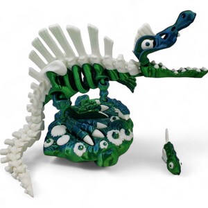 May include: A green and white skeletal dinosaur toy with a blue head and large eyes. The dinosaur is standing on a green and blue base with white eyes. A smaller green and white fish toy is in the foreground.