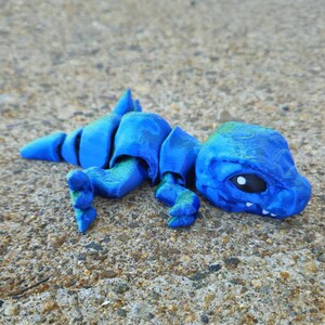 May include: A small, blue, 3D printed dinosaur toy with a segmented body. The dinosaur has a green tint and large, black eyes.