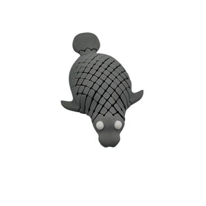 Puede incluir: Una figura de foca gris estilizada con un patr&oacute;n texturizado en forma de escamas. La foca tiene peque&ntilde;os ap&eacute;ndices en forma de aleta y dos ojos redondos blancos. La figura est&aacute; sobre un fondo blanco liso.