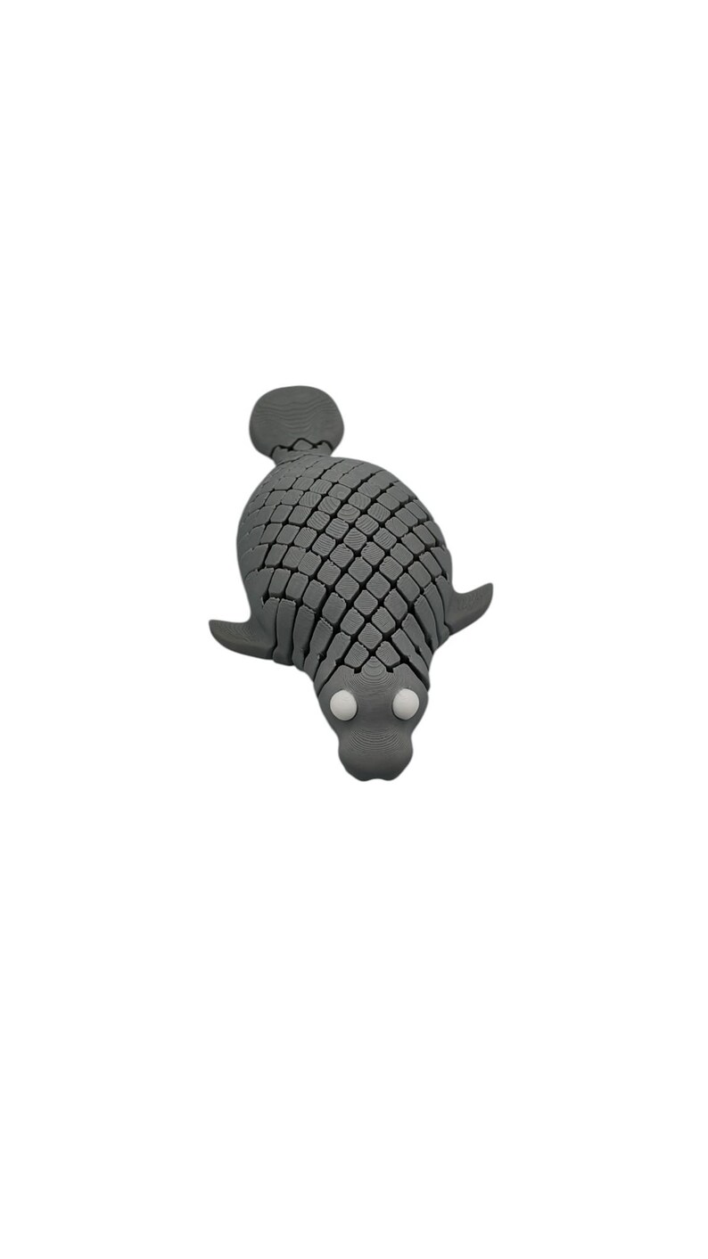Puede incluir: Una figura de foca gris estilizada con un patr&oacute;n texturizado en forma de escamas. La foca tiene peque&ntilde;os ap&eacute;ndices en forma de aleta y dos ojos redondos blancos. La figura est&aacute; sobre un fondo blanco liso.