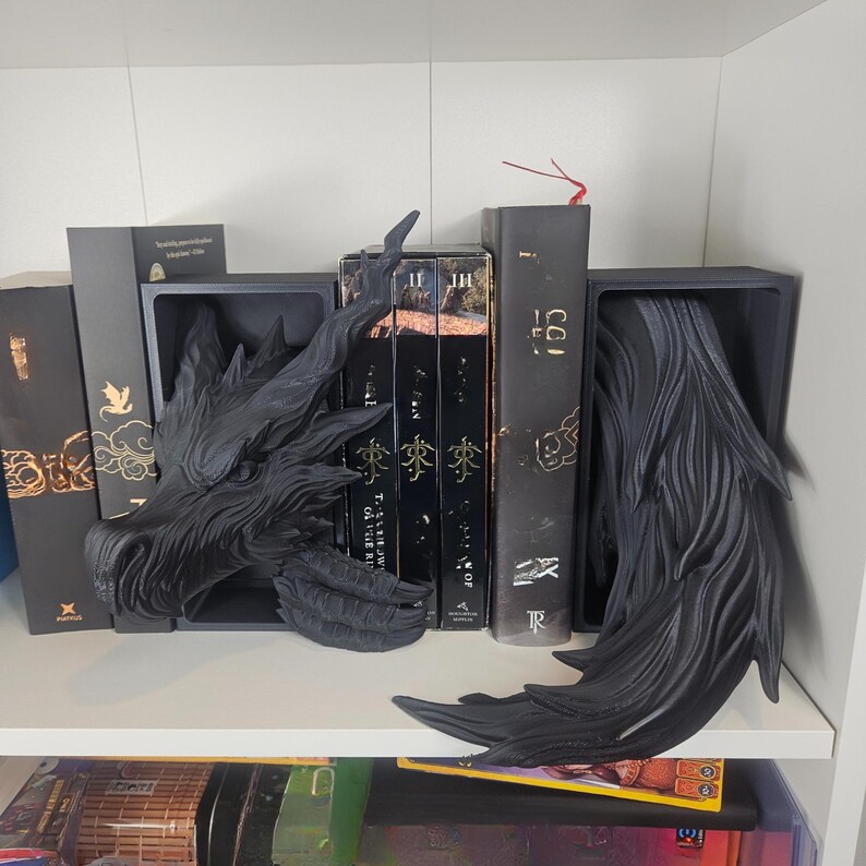 以下が含まれることがあります： 白い棚に置かれた黒いドラゴンの頭と尾のブックエンド。ブックエンドは暗い素材でできており、本を支えるように配置されています。金の文字が入った本がブックエンドの間に見えます。