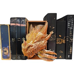 May include: A golden dragon head bookend sits in a wooden box, flanked by several black books with gold lettering. The books have various designs and titles. The dragon head is detailed and textured.