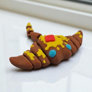 May include: A brown croissant-shaped object with yellow and red accents, and blue dots. The pastry is decorated with what appears to be melted butter and a small red square. The object is on a white surface.