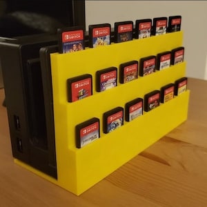 May include: A yellow plastic stand with three shelves for storing Nintendo Switch game cartridges. The stand is holding a variety of game cartridges, including "Civilization VI", "Super Mario Odyssey", and "The Legend of Zelda: Breath of the Wild".