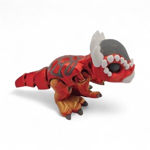 May include: A red and gray 3D-printed dinosaur figurine. The toy dinosaur has a segmented body, with a red body and tail, gray accents, and gold-colored feet. The dinosaur has white horns and teeth, and a gray head.