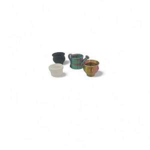 May include: Four miniature ceramic planters in various shapes and colors. Includes a small black pot, a white pot, a green and purple watering can, and a gold-colored pot with pink accents. Perfect for small plants or decorative use.