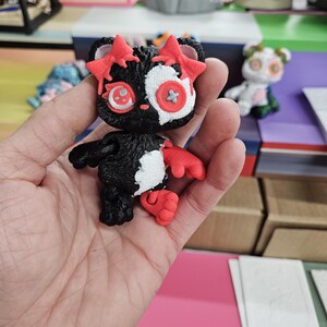 Puede incluir: Una peque&ntilde;a figura en blanco y negro con detalles rojos. La figura tiene detalles en forma de estrella en la cabeza y manos y pies rojos. Los ojos son una mezcla de rojo y blanco, con un detalle en forma de bot&oacute;n en el centro. La figura se sostiene en una mano.