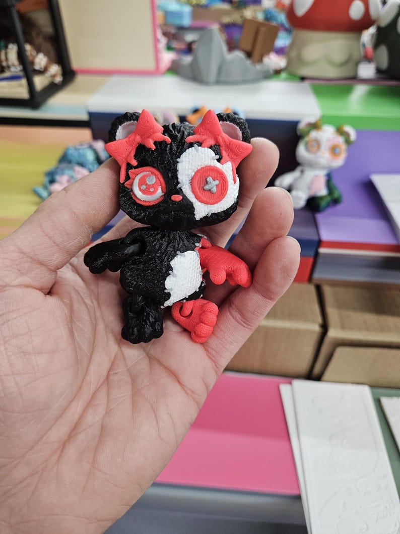 Puede incluir: Una peque&ntilde;a figura en blanco y negro con detalles rojos. La figura tiene detalles en forma de estrella en la cabeza y manos y pies rojos. Los ojos son una mezcla de rojo y blanco, con un detalle en forma de bot&oacute;n en el centro. La figura se sostiene en una mano.