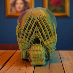 Op de afbeelding: Een 3D-geprinte schedelsculptuur in groen- en goudtinten, met handen die de ogen bedekken. De schedel staat op een houten oppervlak, met ingelijste kunstwerken op de achtergrond. De sculptuur heeft een getextureerd uiterlijk.