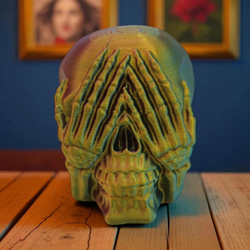 Op de afbeelding: Een 3D-geprinte schedelsculptuur in groen- en goudtinten, met handen die de ogen bedekken. De schedel staat op een houten oppervlak, met ingelijste kunstwerken op de achtergrond. De sculptuur heeft een getextureerd uiterlijk.