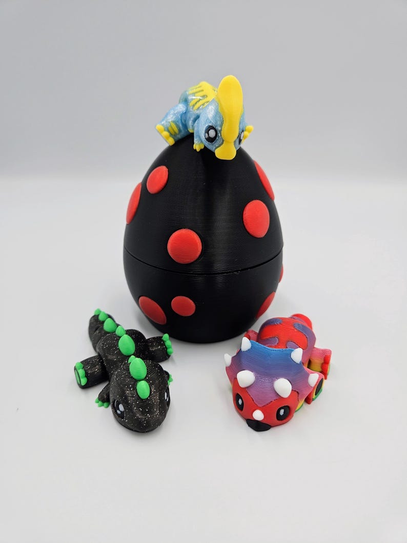May include: Three small dinosaur figurines, one on top of a black egg with red polka dots, one black with green accents, and one multi-colored with white horns.