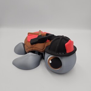 Pode incluir: Estatueta de tartaruga impressa em 3D com tema de bombeiro. A tartaruga tem um corpo cinza, casco marrom e um capacete de bombeiro preto com um detalhe vermelho. Tamb&eacute;m tem uma sela vermelha e ferramentas nas costas.