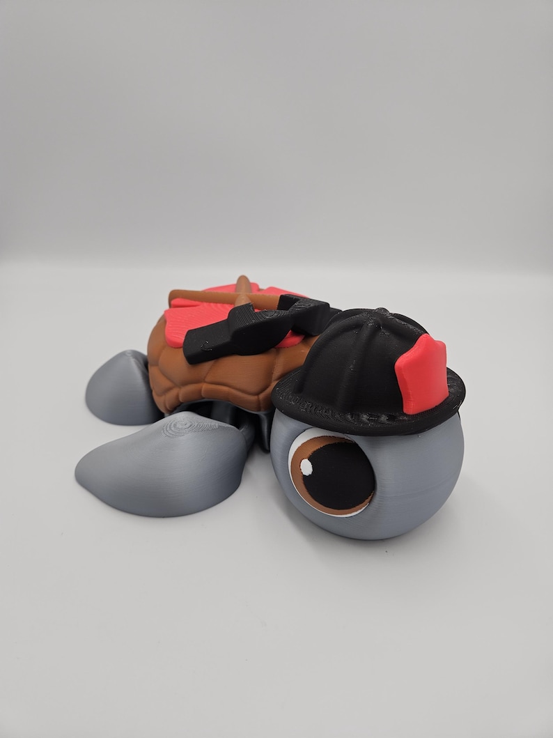 Pode incluir: Estatueta de tartaruga impressa em 3D com tema de bombeiro. A tartaruga tem um corpo cinza, casco marrom e um capacete de bombeiro preto com um detalhe vermelho. Tamb&eacute;m tem uma sela vermelha e ferramentas nas costas.