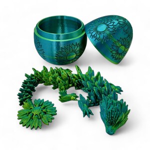 Puede incluir: Una figurita de drag&oacute;n impresa en 3D de color azul verdoso y verde con un dise&ntilde;o de girasol. El drag&oacute;n est&aacute; enroscado y tiene un huevo azul verdoso y verde con un dise&ntilde;o de girasol en la parte superior.