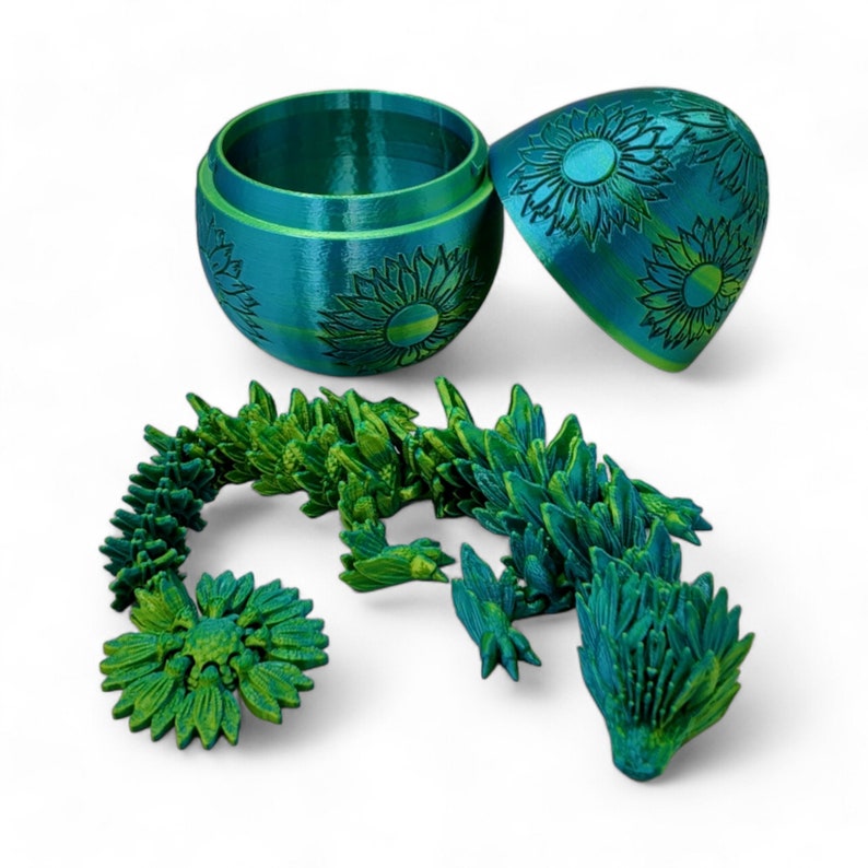 Puede incluir: Una figurita de drag&oacute;n impresa en 3D de color azul verdoso y verde con un dise&ntilde;o de girasol. El drag&oacute;n est&aacute; enroscado y tiene un huevo azul verdoso y verde con un dise&ntilde;o de girasol en la parte superior.