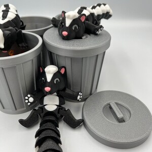 May include: A playful scene featuring 3D-printed skunk figurines in black, white, and pink, posed around gray miniature trash cans. One skunk is lying on a trash can lid. The skunks have articulated bodies and are designed with cartoonish features.
