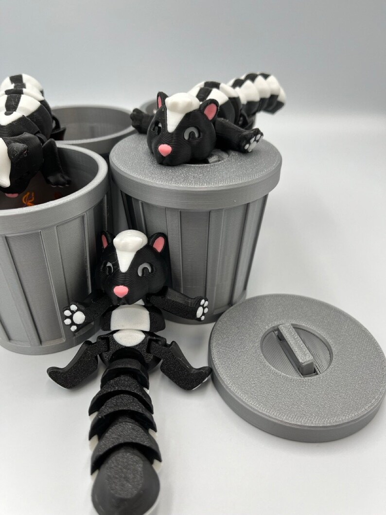 May include: A playful scene featuring 3D-printed skunk figurines in black, white, and pink, posed around gray miniature trash cans. One skunk is lying on a trash can lid. The skunks have articulated bodies and are designed with cartoonish features.