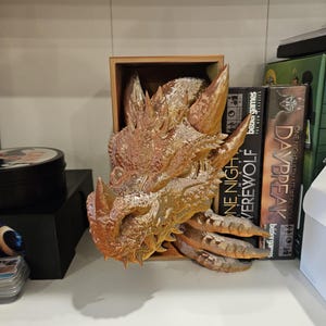 May include: A golden dragon head sculpture with textured scales and sharp teeth. The sculpture is displayed on a wooden shelf with board games in the background.