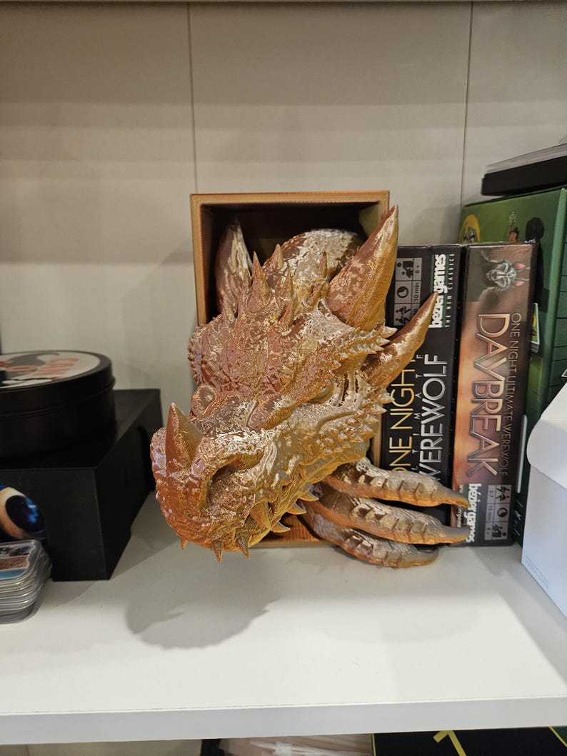 May include: A golden dragon head sculpture with textured scales and sharp teeth. The sculpture is displayed on a wooden shelf with board games in the background.