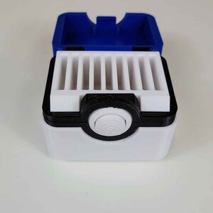 May include: A white and black Pokemon-themed storage box with a blue lid. The box has a circular button on the front and a series of slots inside for holding small items.