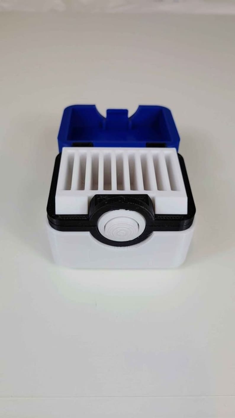 May include: A white and black Pokemon-themed storage box with a blue lid. The box has a circular button on the front and a series of slots inside for holding small items.