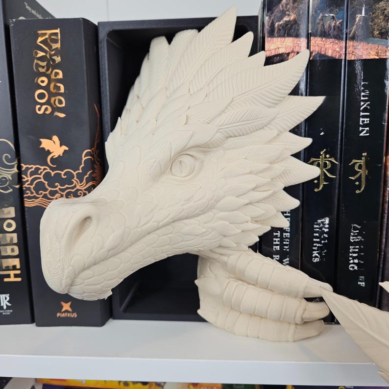 May include: A cream-colored dragon head sculpture with detailed scales and feather-like accents. The dragon's eye is visible, and its clawed hand holds a feather. The sculpture is displayed on a white shelf, with books in the background.