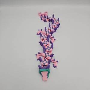 May include: A pink and purple articulated dragon toy with teal accents. The dragon has a segmented body, with a head, tail, and limbs. The dragon is made of plastic and appears to be 3D-printed. The background is plain white.
