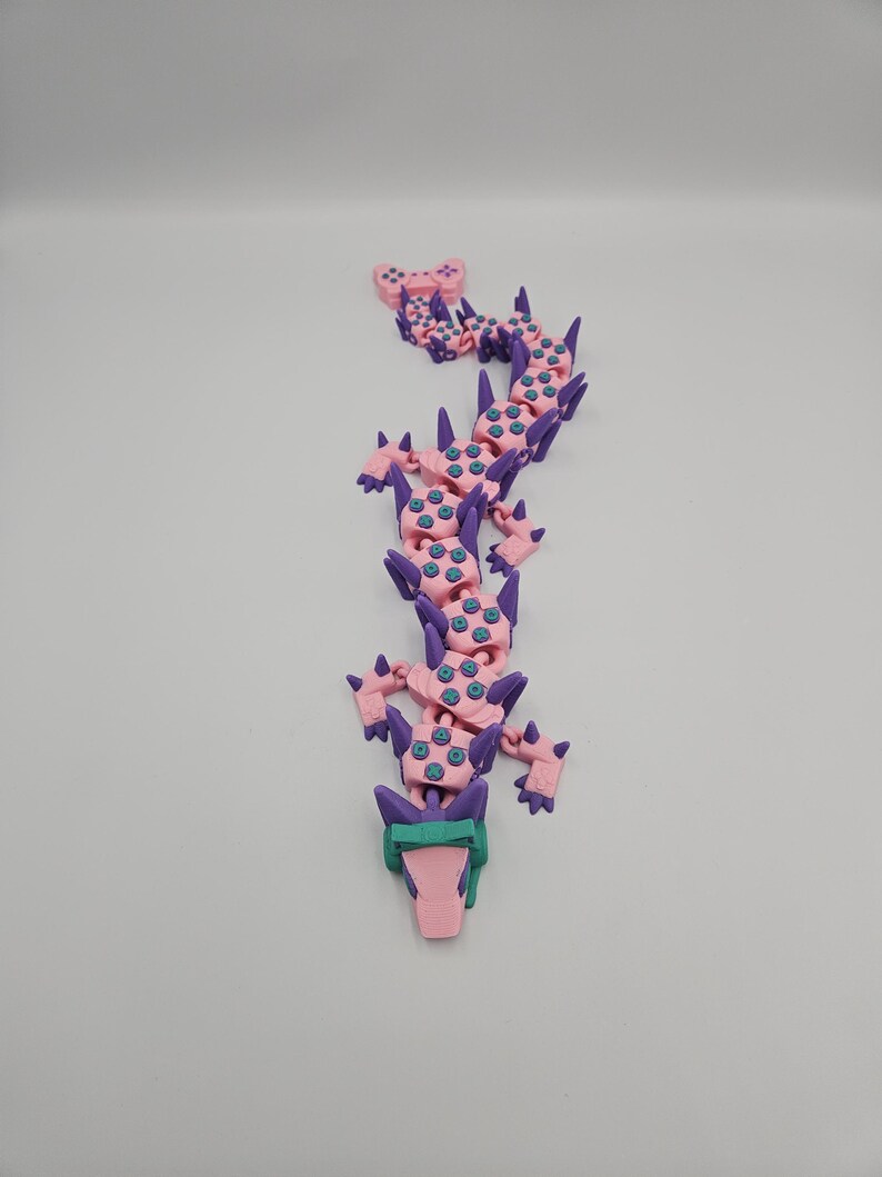 May include: A pink and purple articulated dragon toy with teal accents. The dragon has a segmented body, with a head, tail, and limbs. The dragon is made of plastic and appears to be 3D-printed. The background is plain white.