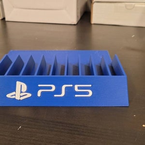 May include: A blue plastic stand with slots for storing video game discs. The stand has the PlayStation logo and the text "PS5" printed on the front.