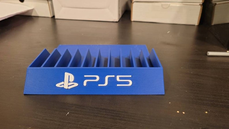 May include: A blue plastic stand with slots for storing video game discs. The stand has the PlayStation logo and the text "PS5" printed on the front.